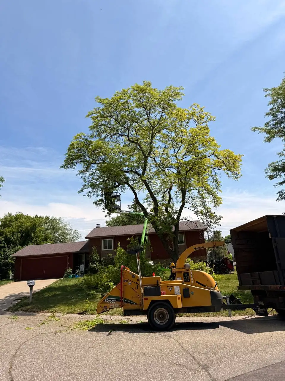 Emergency Tree Removal and Hazard Mitigation Near Structures Tree service expert using a boom lift to safely cut down hazardous tree branches overhanging a residential home during an emergency response call.